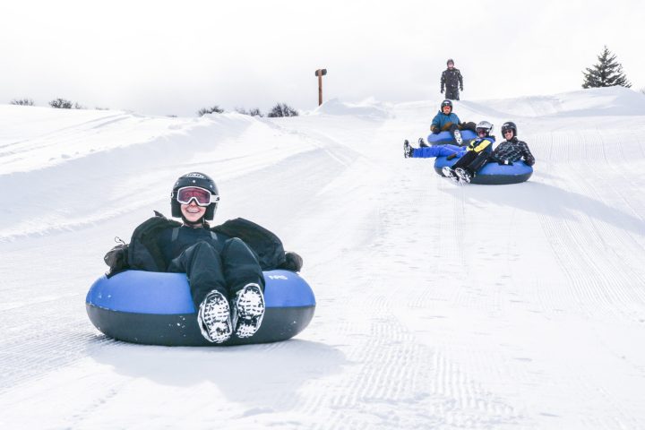 Two people wearing black and helmets sit in snow tubes coming down a freshly groomed trail for snow tubing near Vail, Colorado.