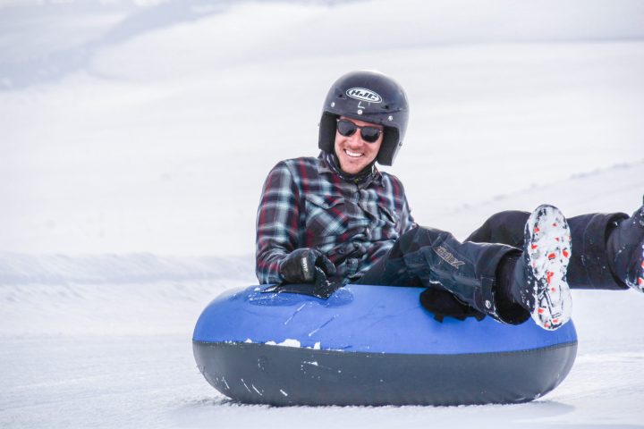 A man sitting on a snow tube with a black helmet and checkered jacket, smiling as he tubes down the groomed trails at the Excursion Resort in Colorado.