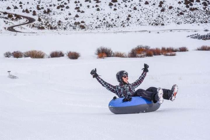 a man on a blue and black individual snowtube has his hands up with a smile on his face showing his excitement while snow tubing down the trail near Vail.