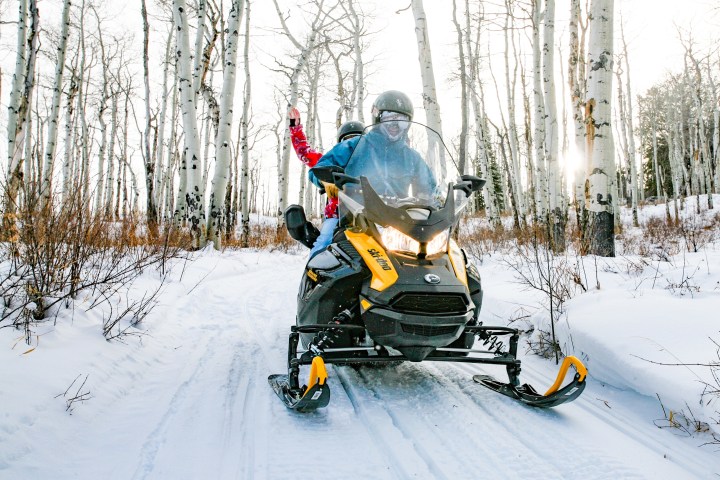 Two people riding a yellow snowmobile through a snowy forest of tall birch trees.