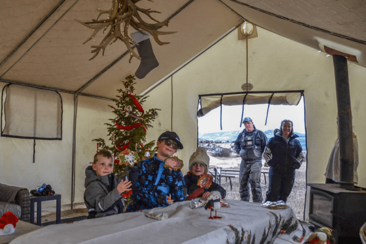 Children in winter clothes indoors with Christmas decorations, adults at the tent entrance, snowy landscape outside.